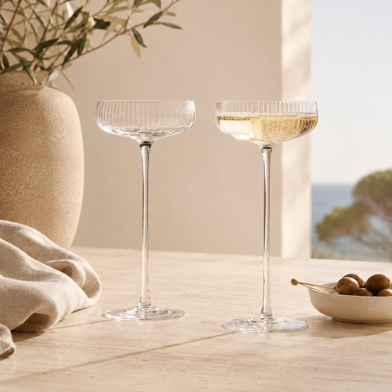 Two clear ribbed-stemmed coupe glasses on a light stone table, with one glass partly filled with pale champagne. A beige vase with branches, a folded beige cloth, and a small dish of olives sit nearby, with a softly blurred outdoor scene in the background.