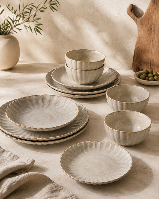 A coordinated set of ceramic tableware in a warm off-white, textured glaze with scalloped edges, arranged on a beige tablecloth with natural shadows. The set includes plates and bowls in various sizes, styled with a linen napkin, a small vase with greenery, and a wooden cutting board in the background.