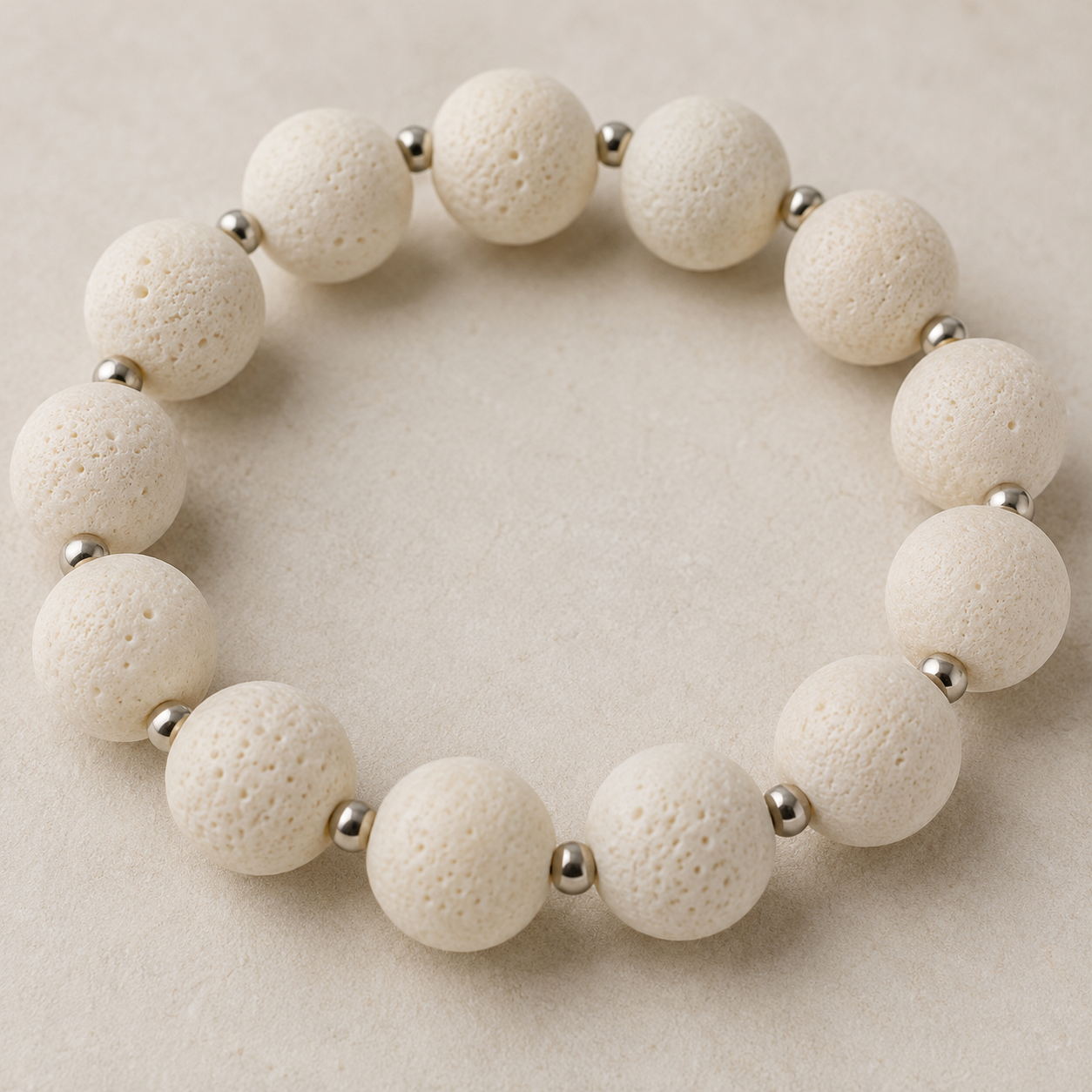 A beaded stretch bracelet made of matte off-white porous lava stone beads separated by small polished metallic silver spacer beads, displayed on a neutral beige textured background.