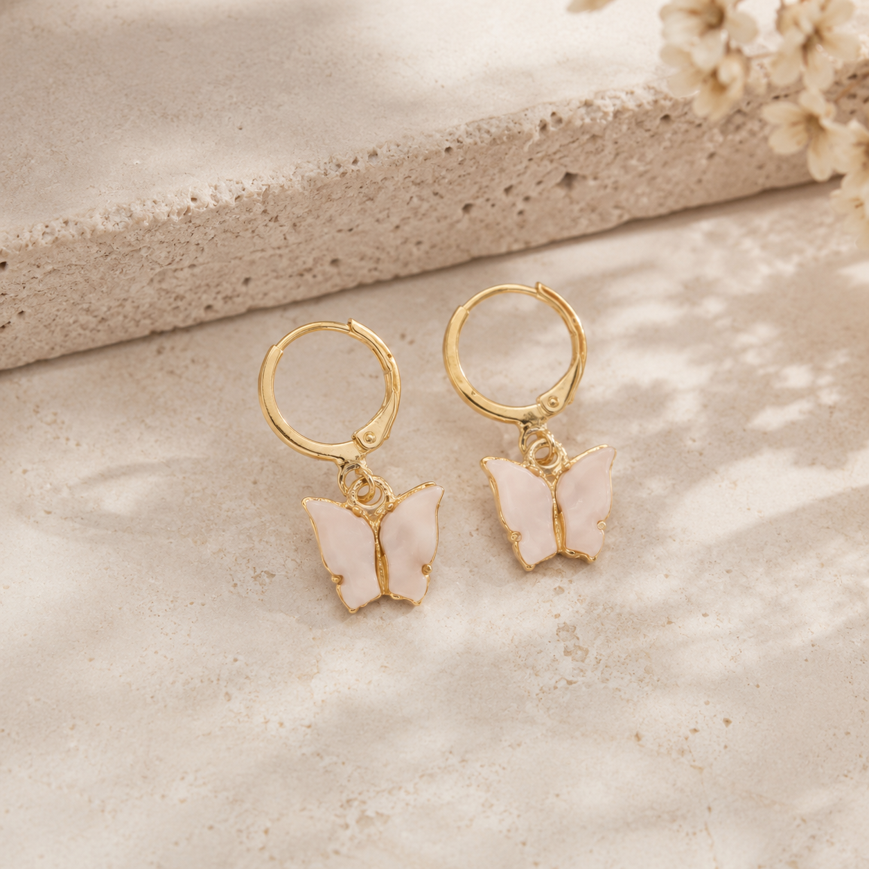 A pair of gold hoop earrings with dangling butterfly charms in pale pink enamel, displayed on a textured beige stone surface.