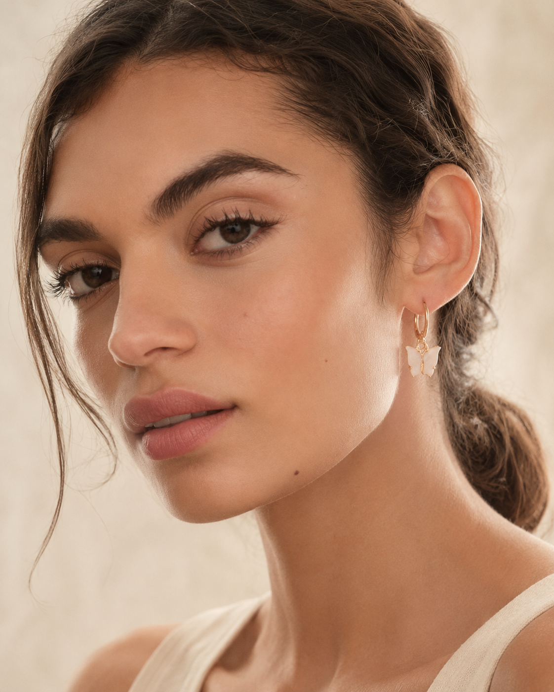 A close-up of a woman wearing a delicate gold hoop earring with a small white butterfly charm, set against a soft beige background.