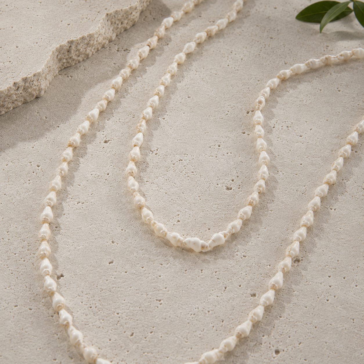 A long strand necklace made of small irregular shell-like beads in an off-white cream color, draped on a textured light beige surface with a piece of coral and green leaves in the corner.