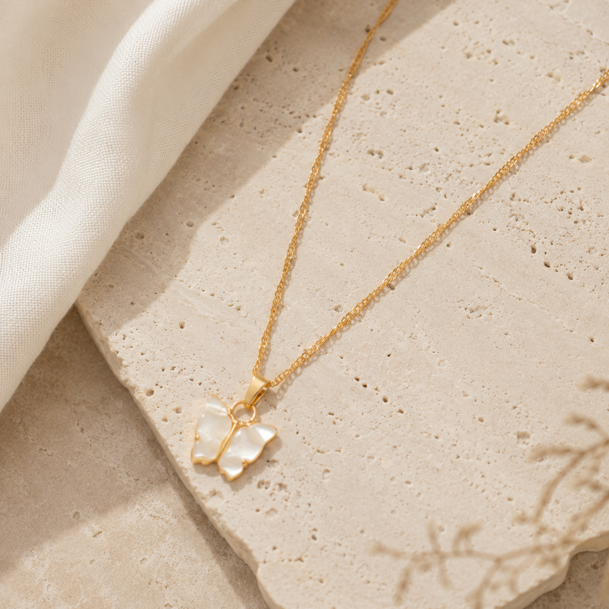 A delicate gold chain necklace with a butterfly pendant made of iridescent mother-of-pearl, styled on a textured beige stone slab with a soft white fabric drape.