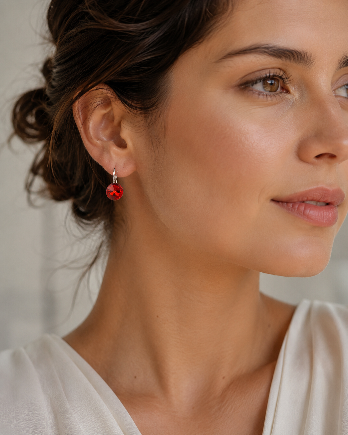 A close-up of a woman wearing a small red dangling drop earring with a silver hook, shown in profile against a soft light background.