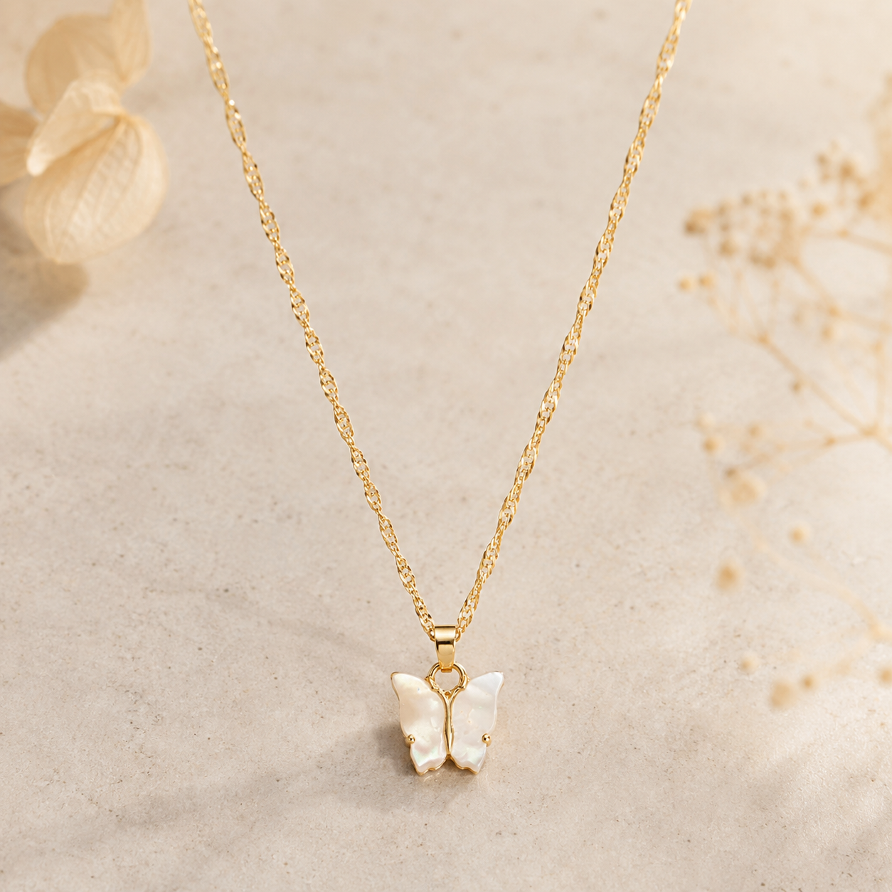A delicate gold chain necklace with a butterfly pendant made of iridescent white mother-of-pearl, displayed on a textured beige surface with soft dried floral accents.